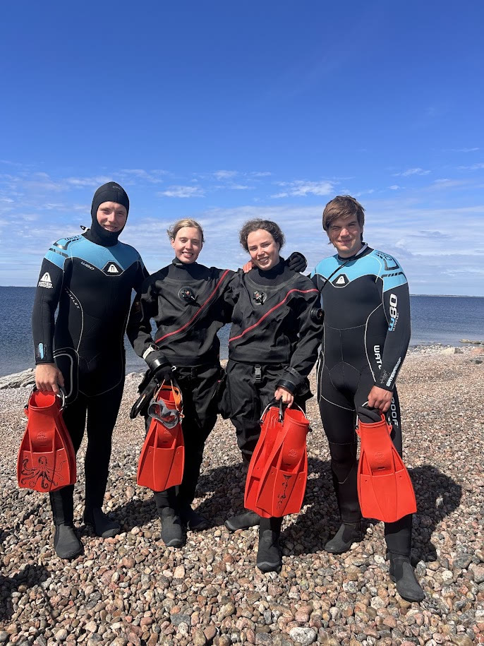 Four BaltiskaBönderna divers in wetsuits holding their fins on the shore
